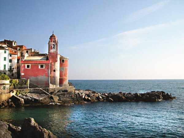 Un'immagine del borgo di Tellaro. Sullo sfondo, in alto, il cielo blu. In basso il mare azzurro. Al centro, su uno scoglio, la chiesa di San Giorgio di colore rosso.